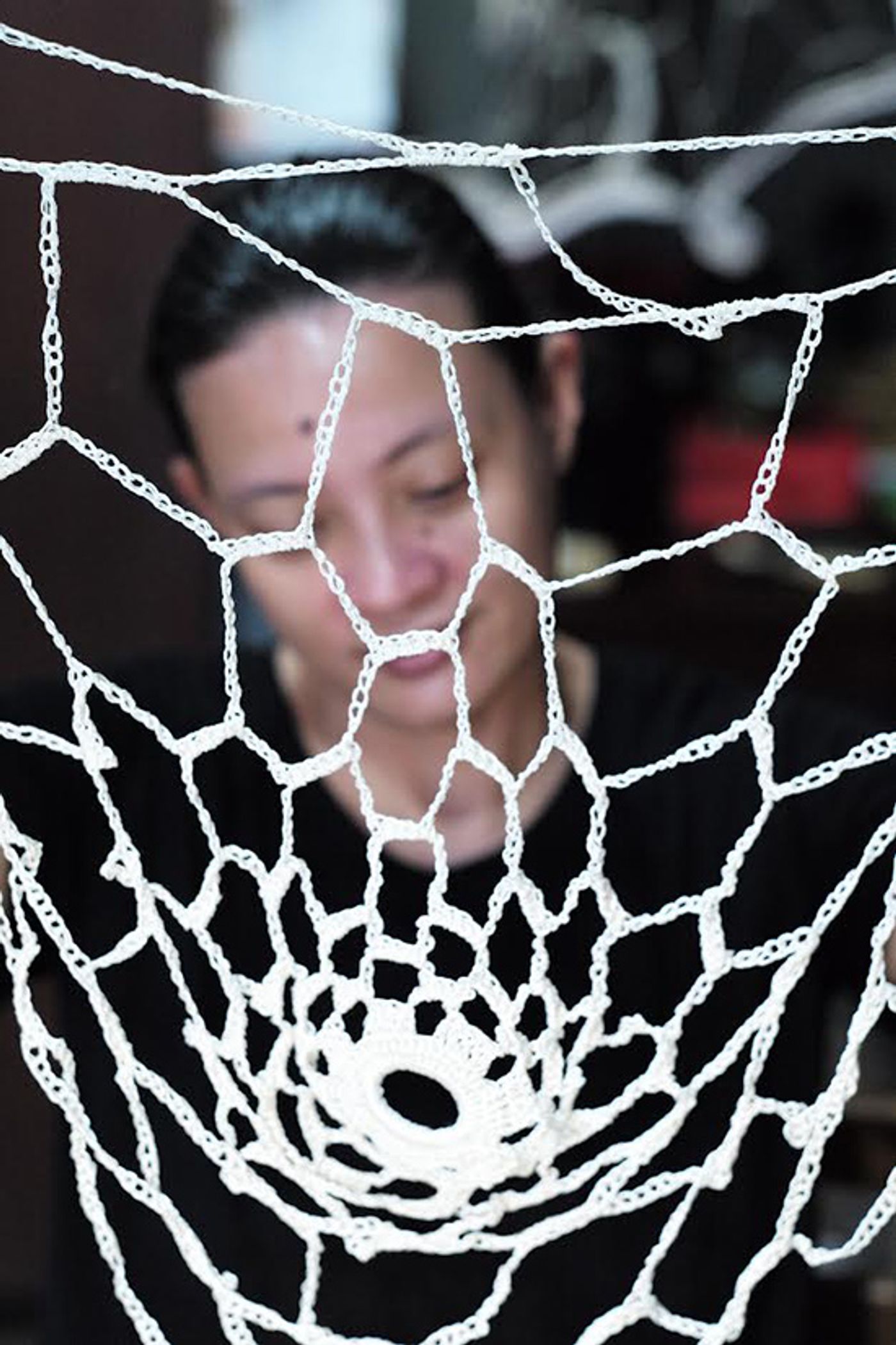 A white crocheted web in sharp focus, artist Aze Ong in black softly blurred behind it