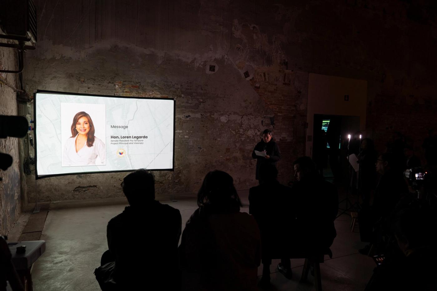 In a dark exhibition hall, Senator Loren Legarda’s portrait and a “Message” slide glow on a screen as a small audience sits in silhouette