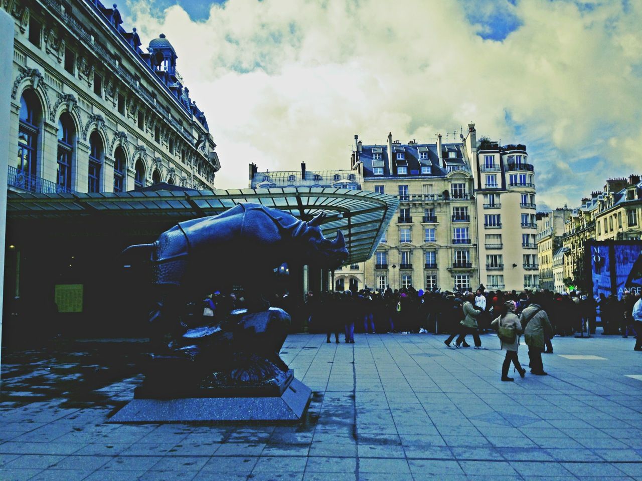 Bronze rhinoceros sculpture outside a glass canopied museum entrance where visitors queue under Haussmann facades