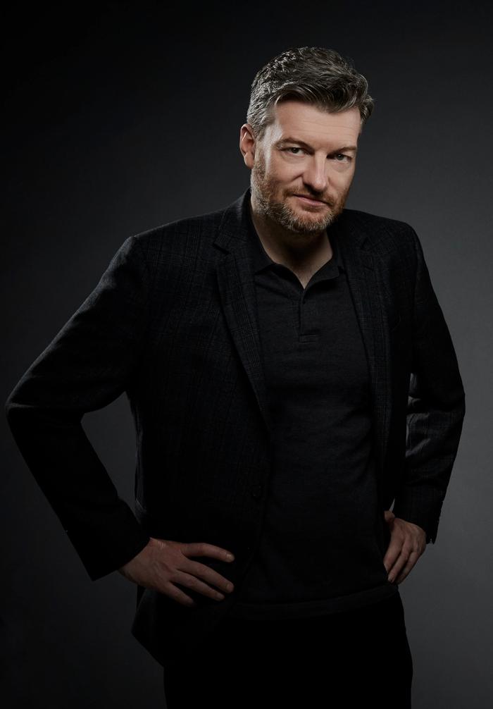 Black Mirror creator Charlie Brooker in a dark blazer over a charcoal polo, hands on hips with a wry half-smile, lit against a grey studio backdrop