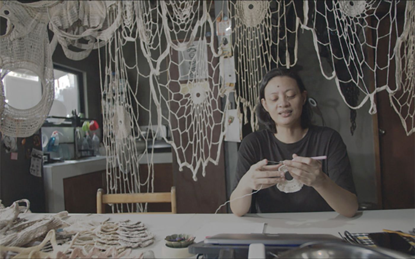 Aze Ong crocheting at a work table in her studio, the walls draped with webs of white crochet