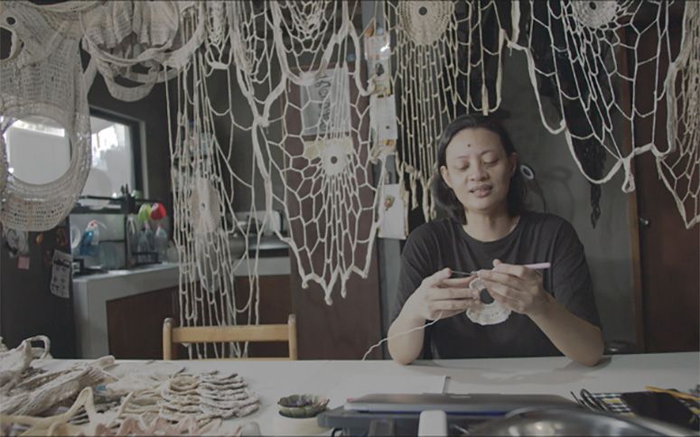 Aze Ong crocheting at a work table in her studio, the walls draped with webs of white crochet