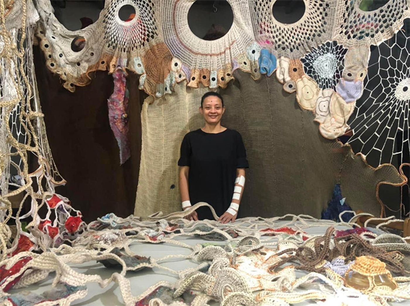 Aze Ong surrounded by her crochet work: looping ropes and eyelike discs spreading across wall and table