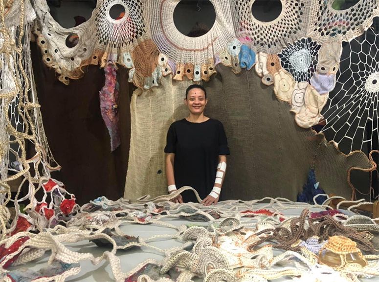 Aze Ong surrounded by her crochet work: looping ropes and eyelike discs spreading across wall and table