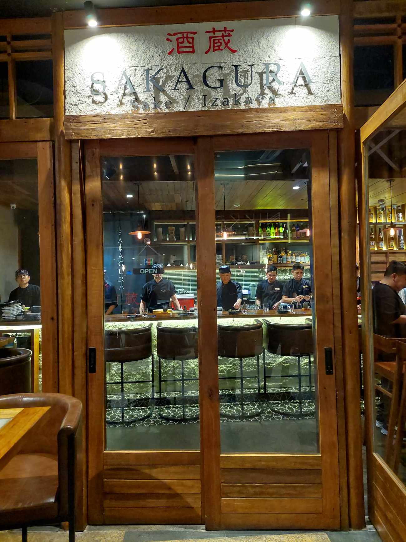 The wood-framed glass entrance of Sakagura Sake and Izakaya, its kanji sign above chefs working the bar counter inside
