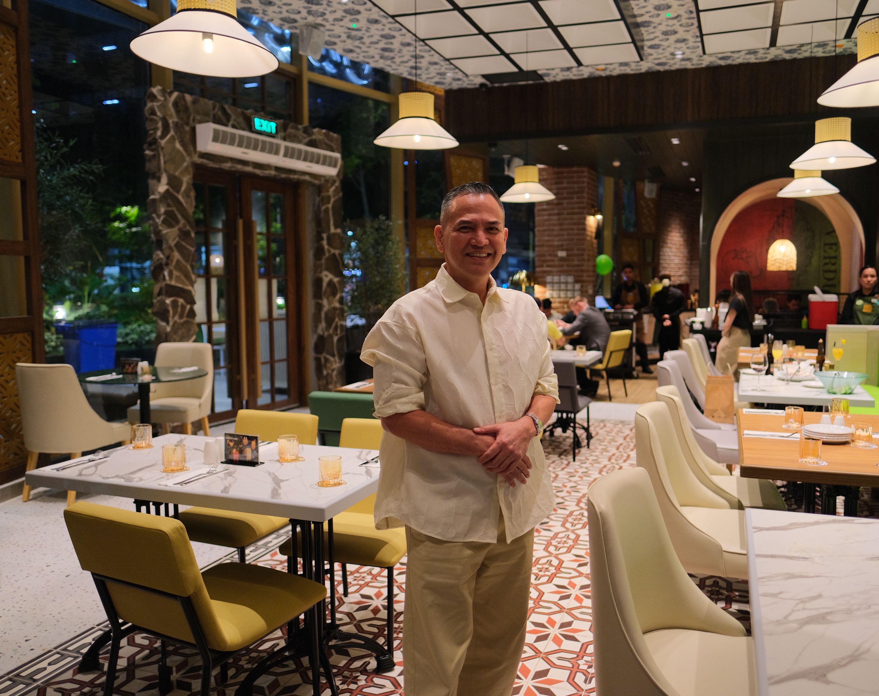 A smiling man in cream stands among marble tables, yellow chairs and patterned tile at Siklab+