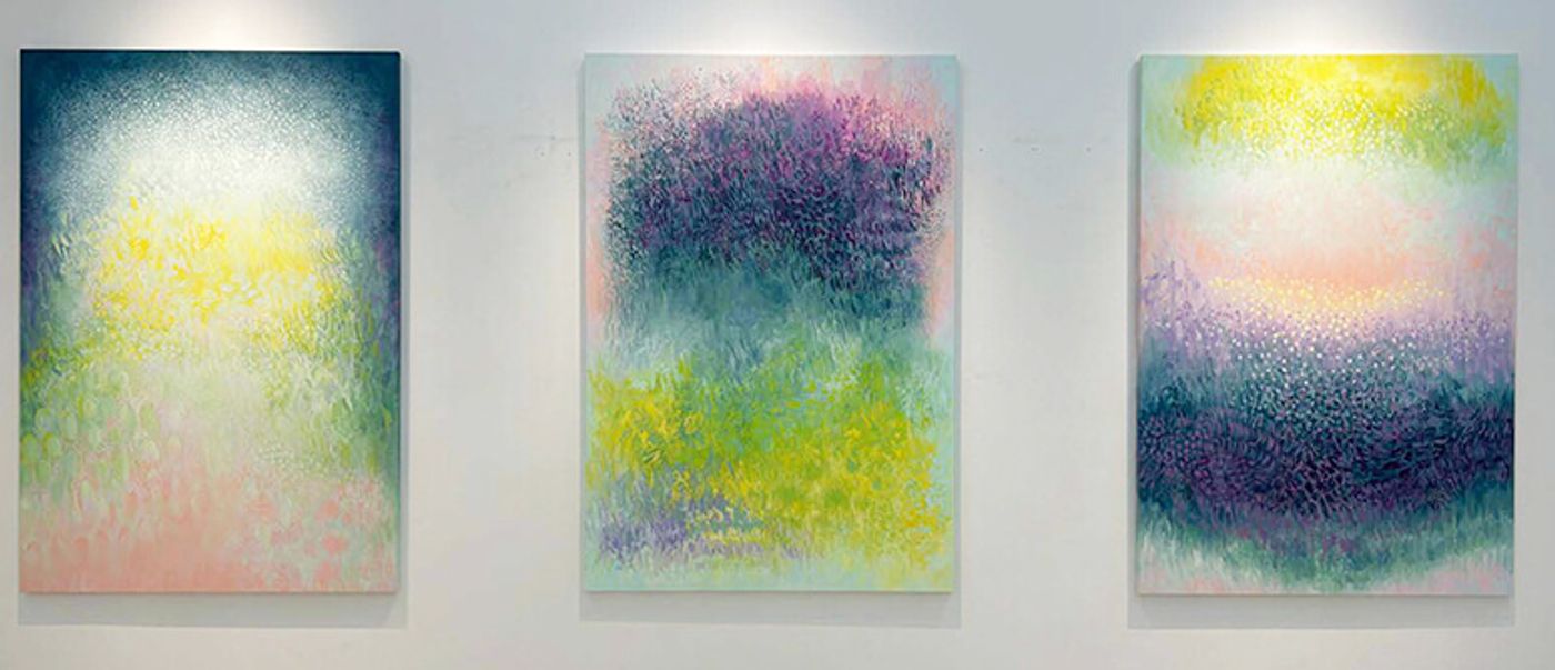 Three large abstract canvases hung side by side on a white gallery wall, each layered with soft mark-making in teal, yellow, pink and purple that transitions from dark to light
