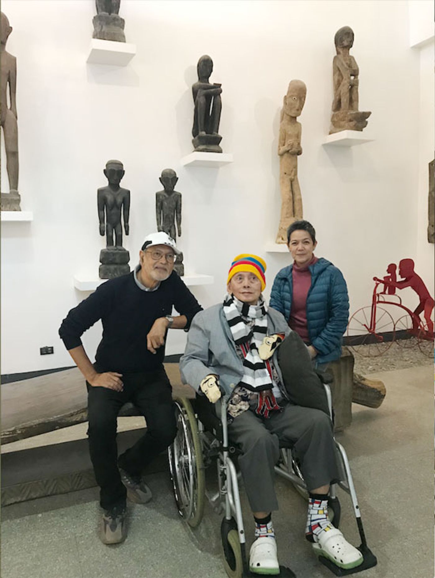 David Medalla in a wheelchair flanked by BenCab and Boots Herrera beneath shelves of carved wooden figures