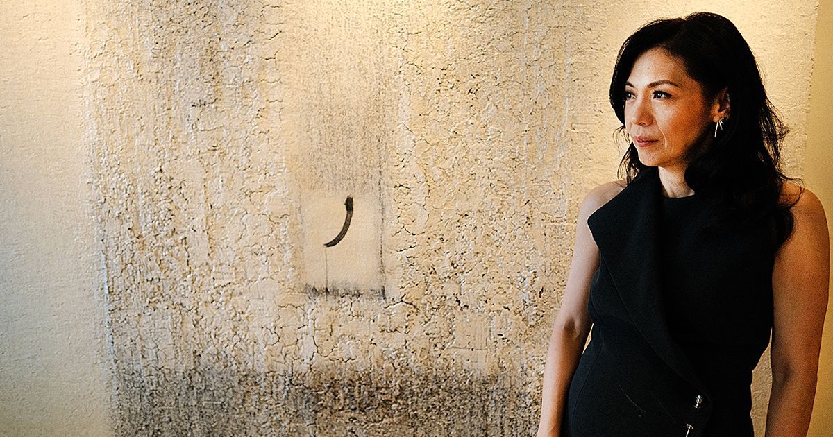 Kaye Tinga, president of the Cultural Center of the Philippines, standing in a black top against a textured wall with a minimalist abstract painting