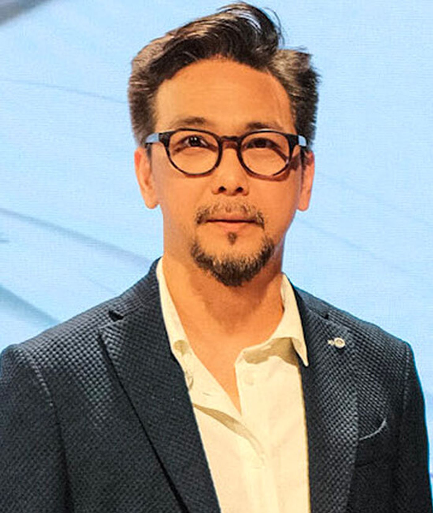 Stanley Ruiz in a textured navy blazer, round black glasses and a trimmed goatee against a pale blue backdrop