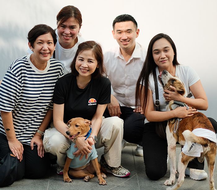 Five Bistro Group executives crouch together with two rescued dogs, a small tan dog and a larger white-and-brown one, against a pale wall