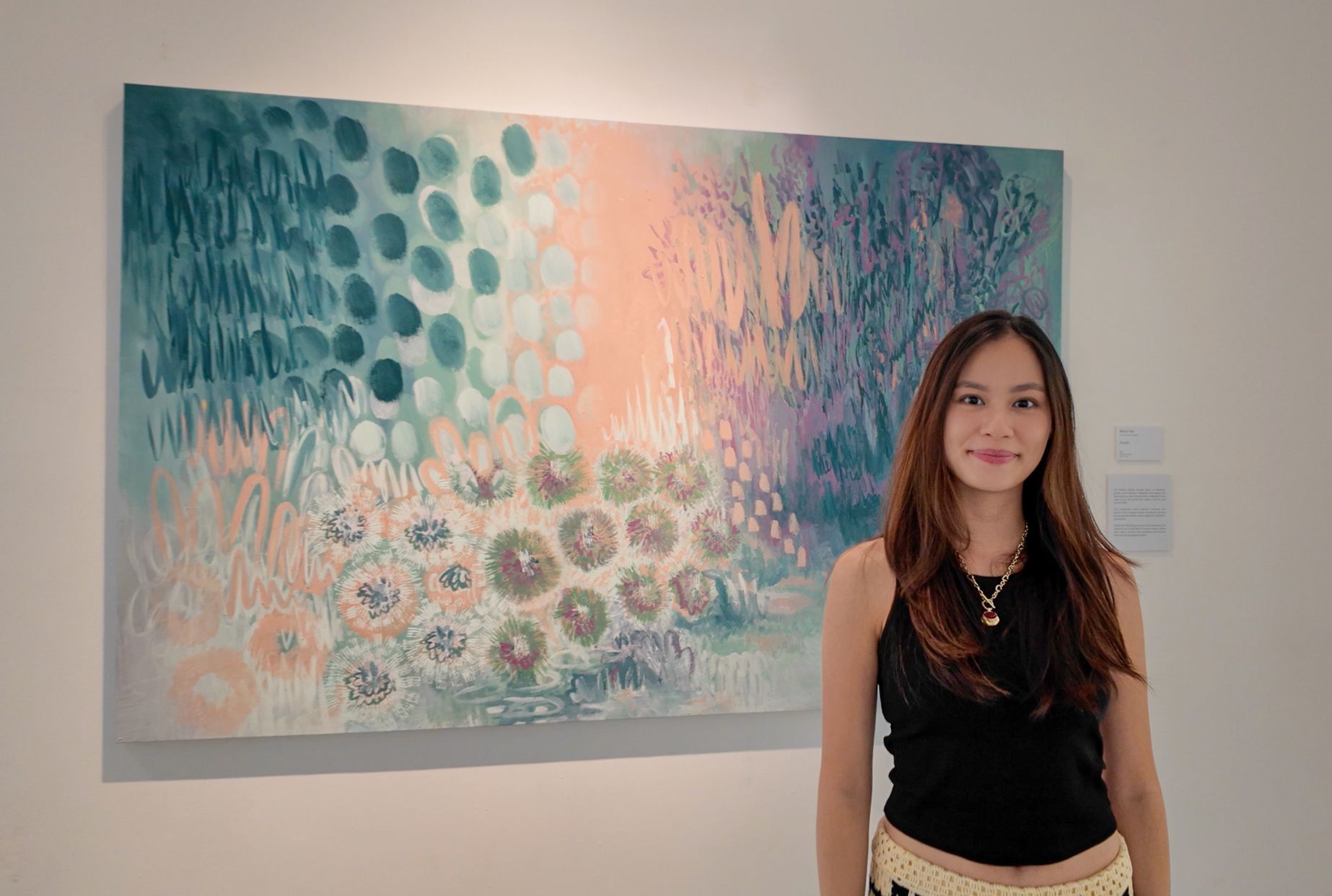 Maria Tan in a black top beside a large abstract painting of teal dots, peach light and flower-like bursts
