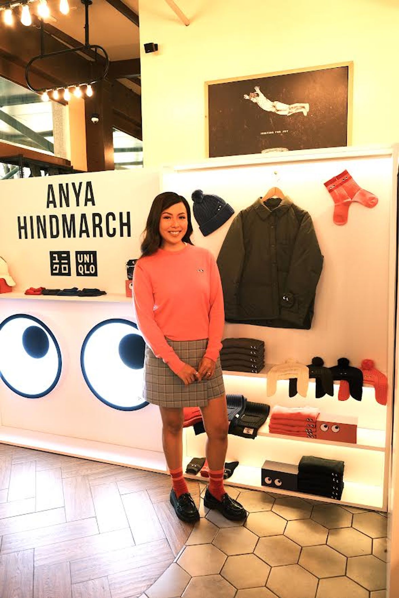 Reiny Vergara in a coral Uniqlo x Anya Hindmarch sweater and a checked skirt, standing beside a store display headed Anya Hindmarch with the Uniqlo logo, hung with a bobble hat, an olive jacket and red socks above shelves of folded knitwear, next to a white panel painted with two large cartoon eyes