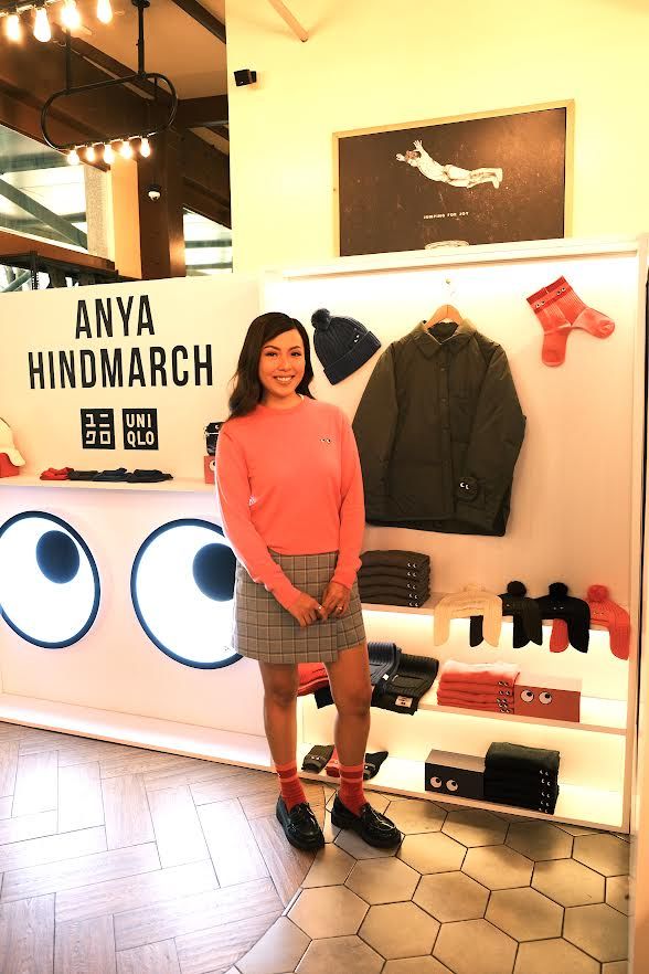 Reiny Vergara in a coral Uniqlo x Anya Hindmarch sweater and a checked skirt, standing beside a store display headed Anya Hindmarch with the Uniqlo logo, hung with a bobble hat, an olive jacket and red socks above shelves of folded knitwear, next to a white panel painted with two large cartoon eyes