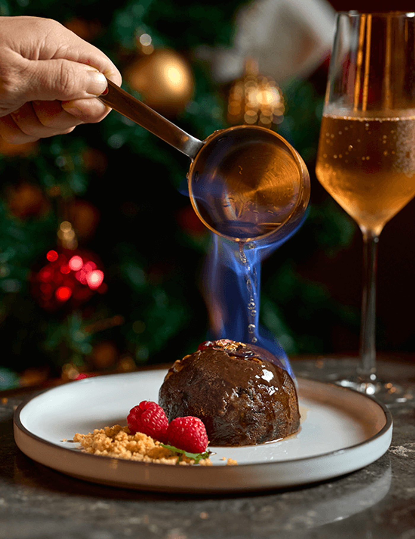 Flaming brandy poured from a copper pan over a dark Christmas pudding with raspberries