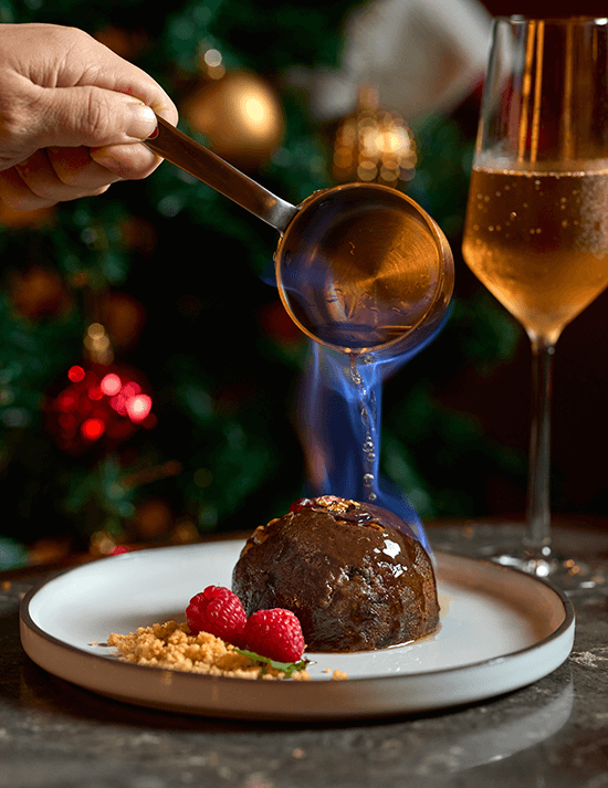 Flaming brandy poured from a copper pan over a dark Christmas pudding with raspberries