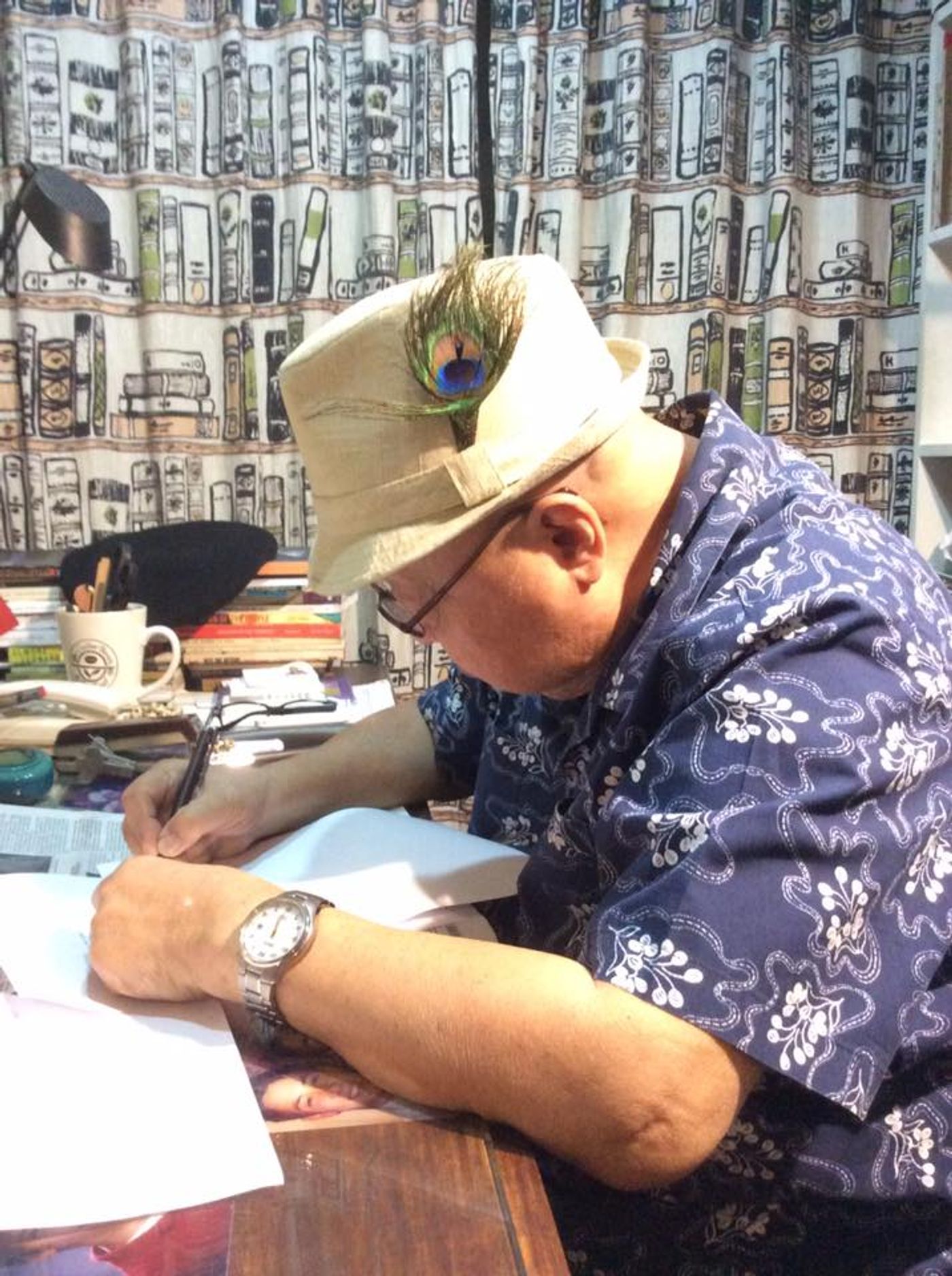 F. Sionil José in a peacock-feathered hat and blue floral shirt signs a book at his cluttered desk