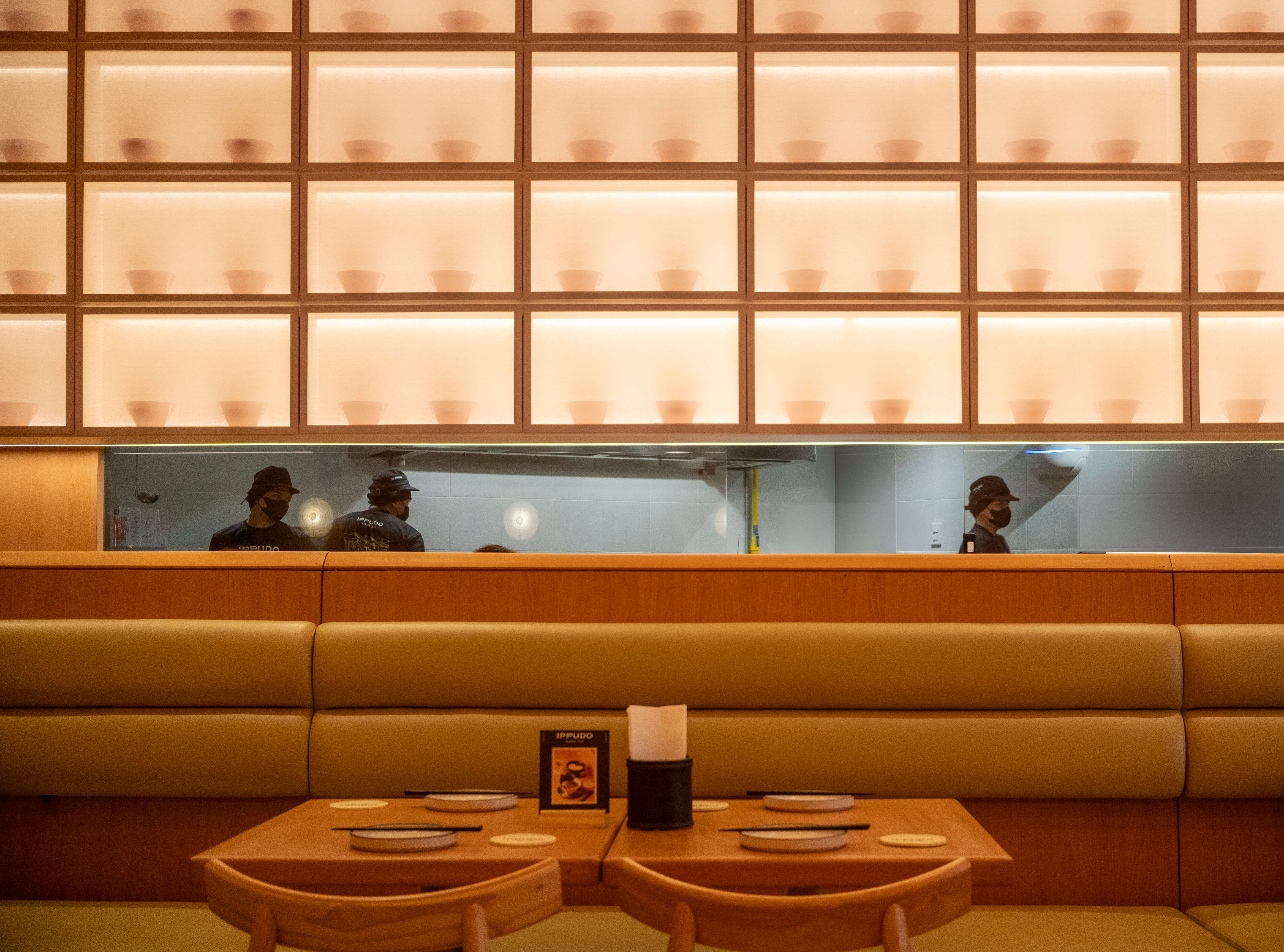 The warm wood interior of Ippudo, a wall of backlit ramen-bowl niches above banquette seating, staff in the open kitchen behind