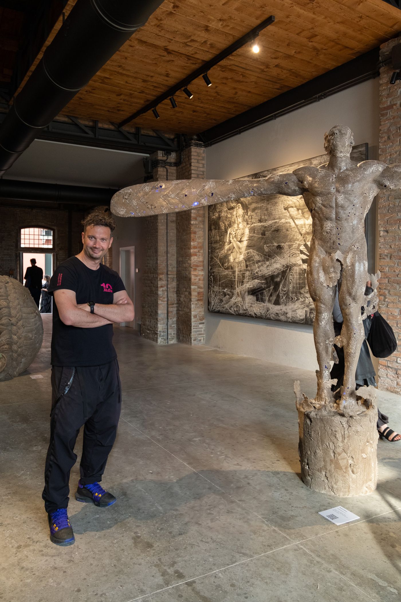 Carlo Maiolini stands arms crossed beside “Consecration,” a pale sculpted figure bearing a long paddle across its shoulders