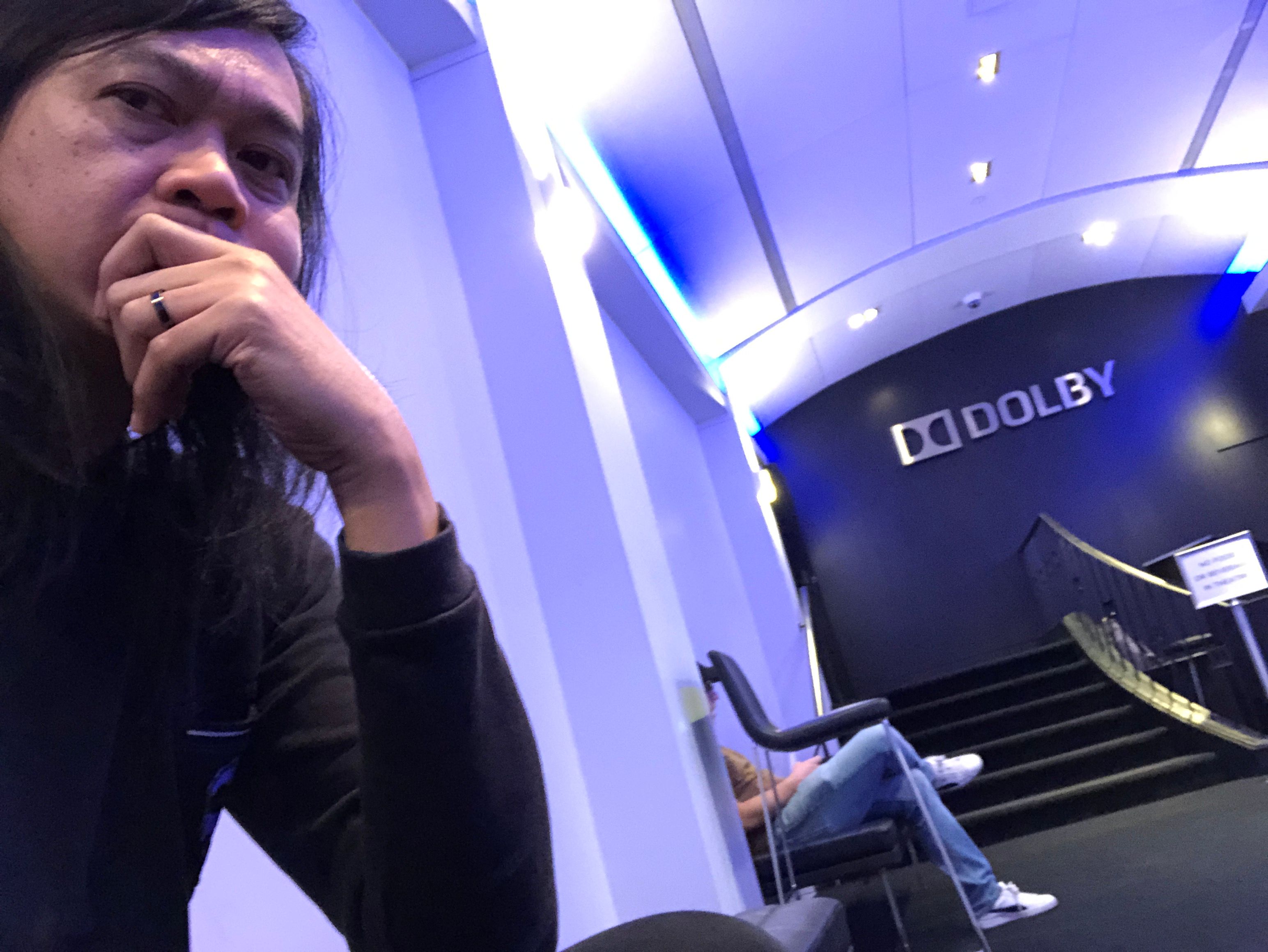 Selfie of a long-haired man looking pensive in a purple-lit lobby, a Dolby sign and spiral stair behind him