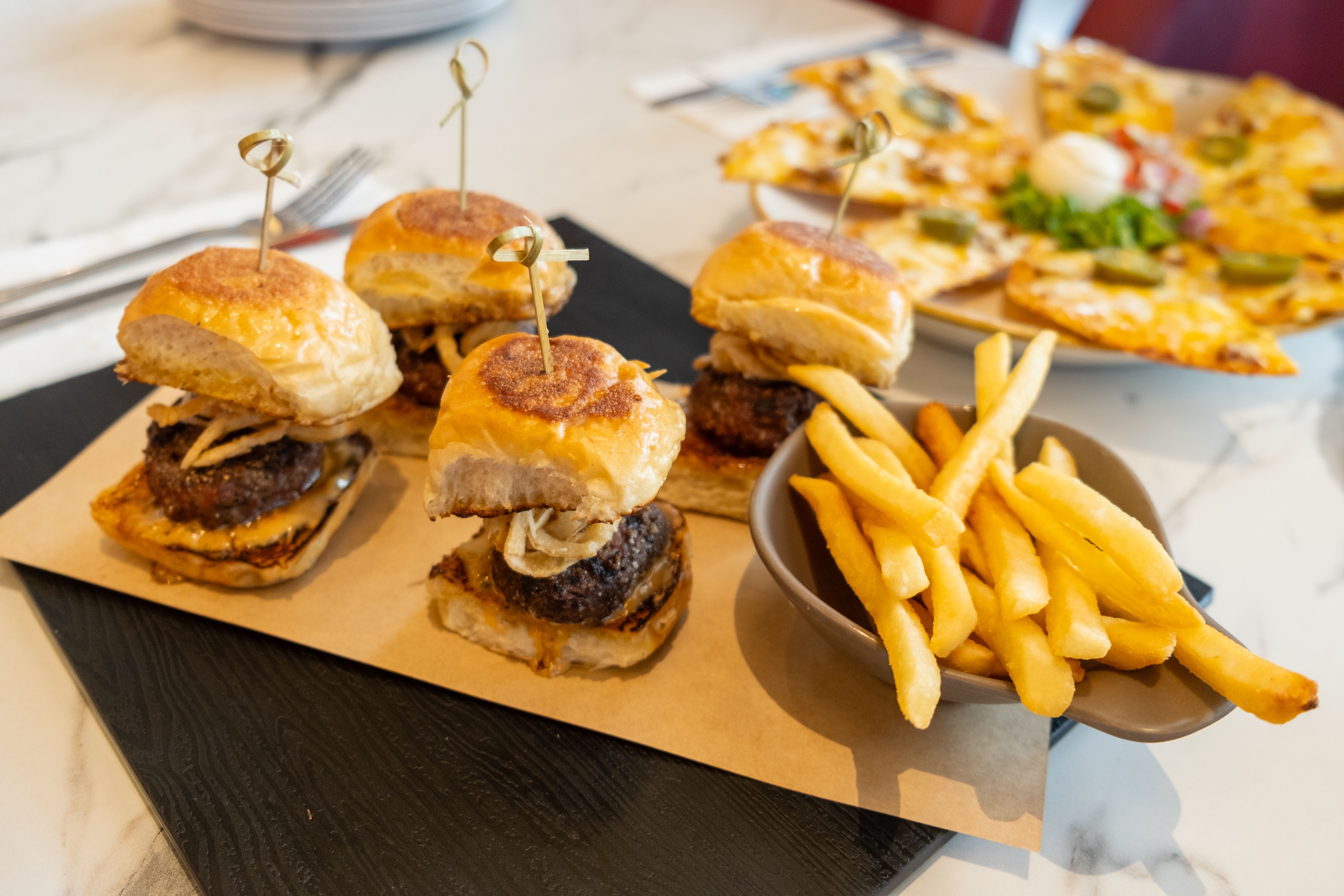 Skewered beef sliders lined up on a wooden board beside a bowl of golden fries