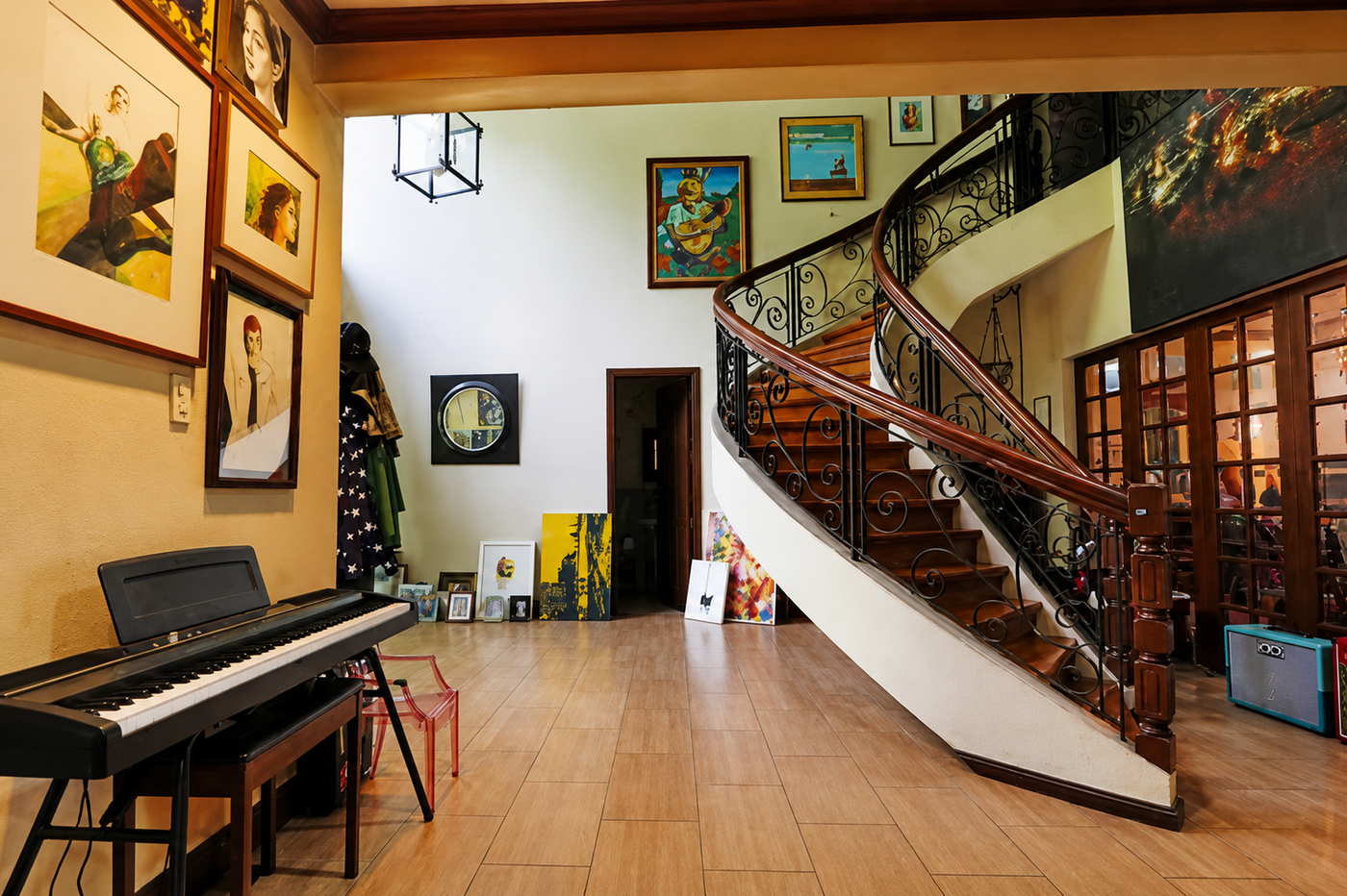 A digital piano and leaning canvases at one end of a high-ceilinged living room, with a curving wrought-iron staircase and walls hung with paintings