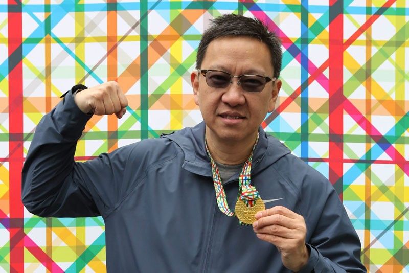 Emerson Yao raises a fist and holds up a marathon finisher medal, in a blue running jacket and sunglasses against a bright criss-crossed backdrop