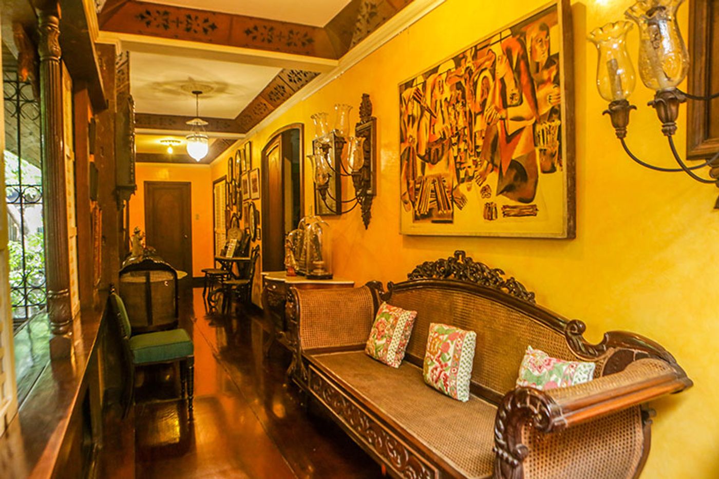 A yellow hallway hung with a large modernist painting, glass hurricane lamps in iron sconces and a caned narra bench with printed cushions