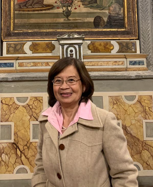 An older woman in glasses, a pink collared shirt and a tan coat smiles before an ornate chapel wall with a gilt-framed painting and faux-marble panels