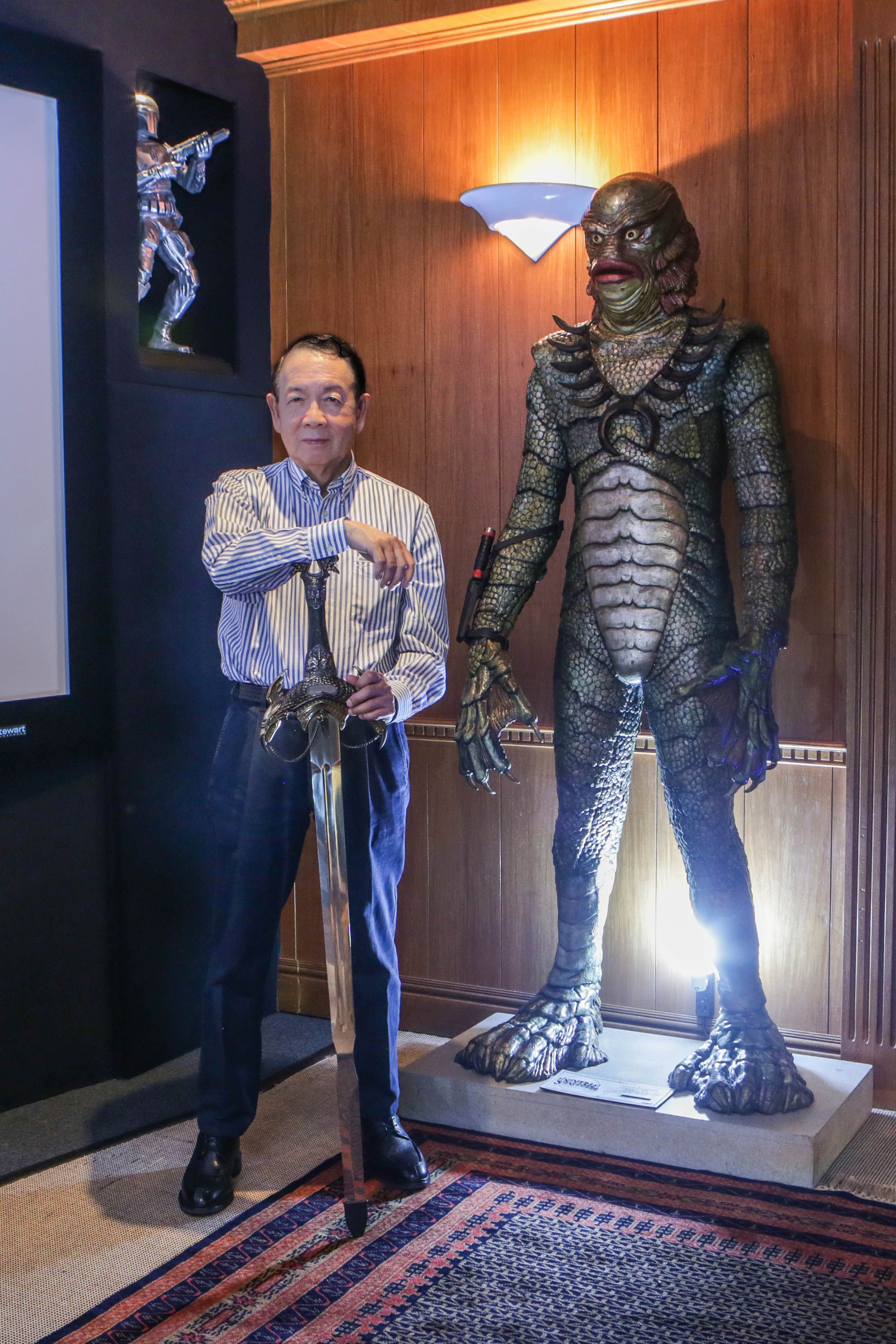 Jun Lopez in a striped shirt leaning on a broadsword beside a life-size Creature from the Black Lagoon figure, a Boba Fett statue lit in a niche behind him