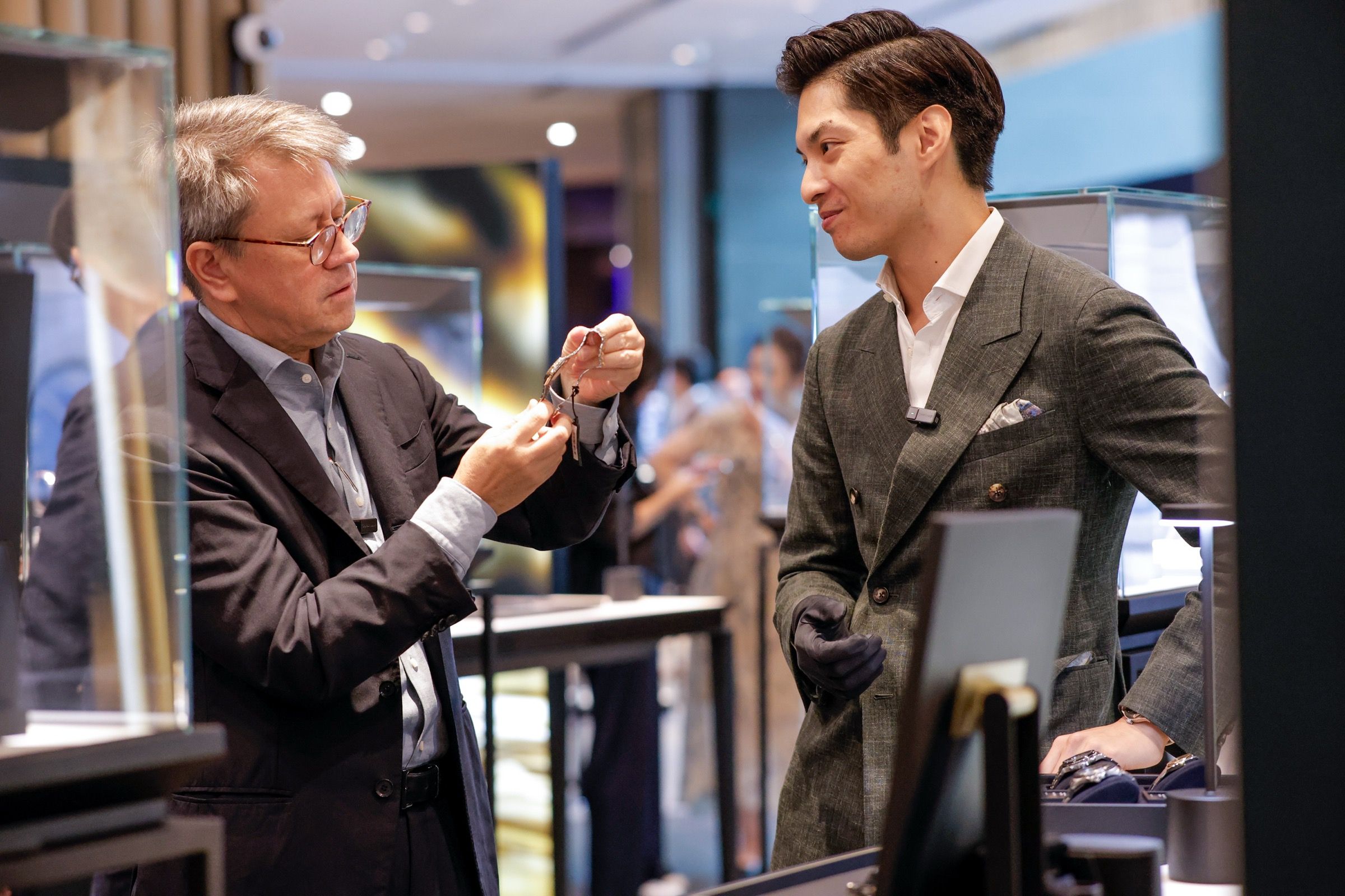 A watchmaker in glasses examining a mechanical watch held in hand while speaking with a visitor in a double-breasted suit at Lucerne Watch Week