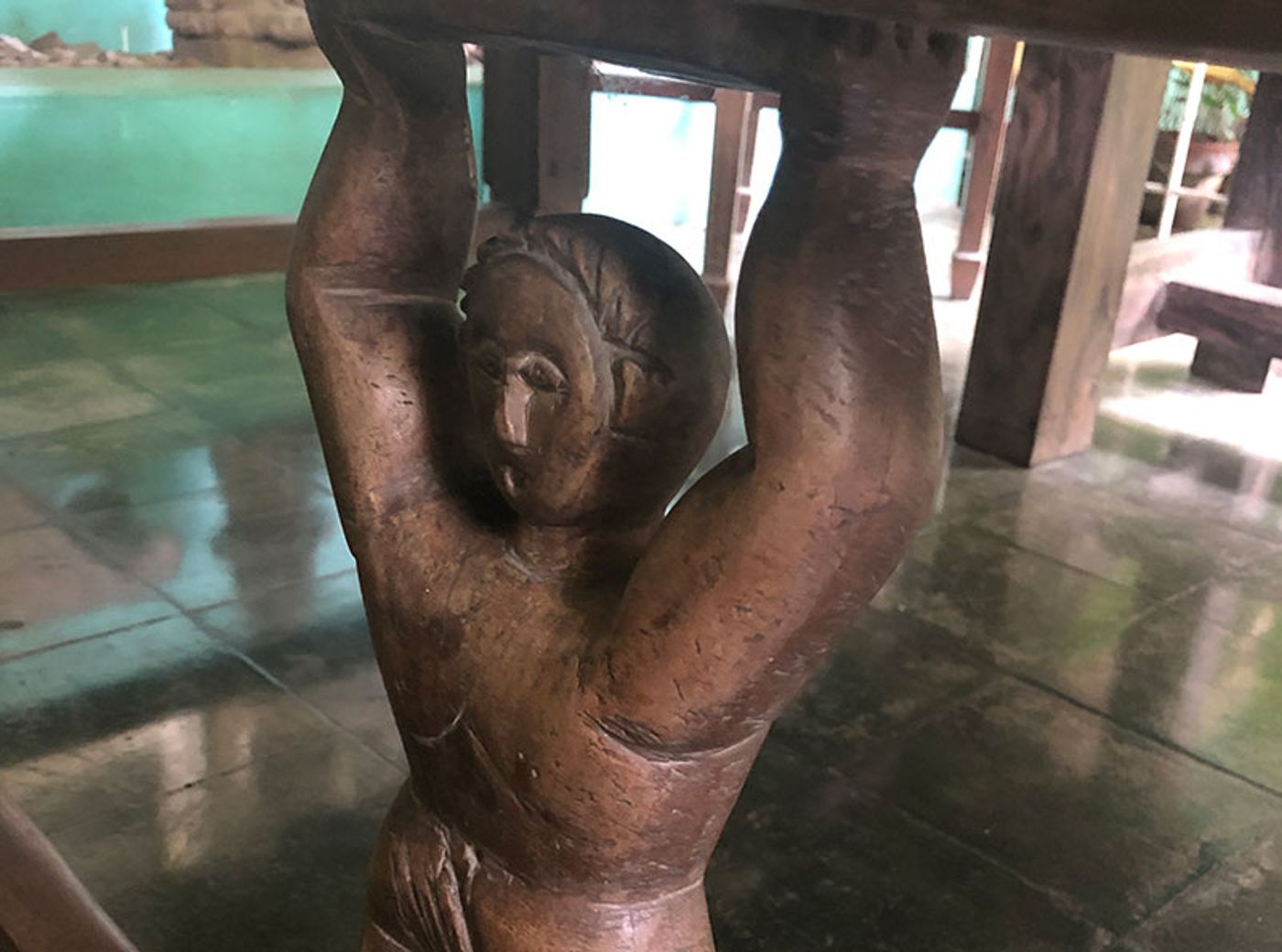 A carved wooden figure with both arms raised, holding up the top of a round table on a tiled floor