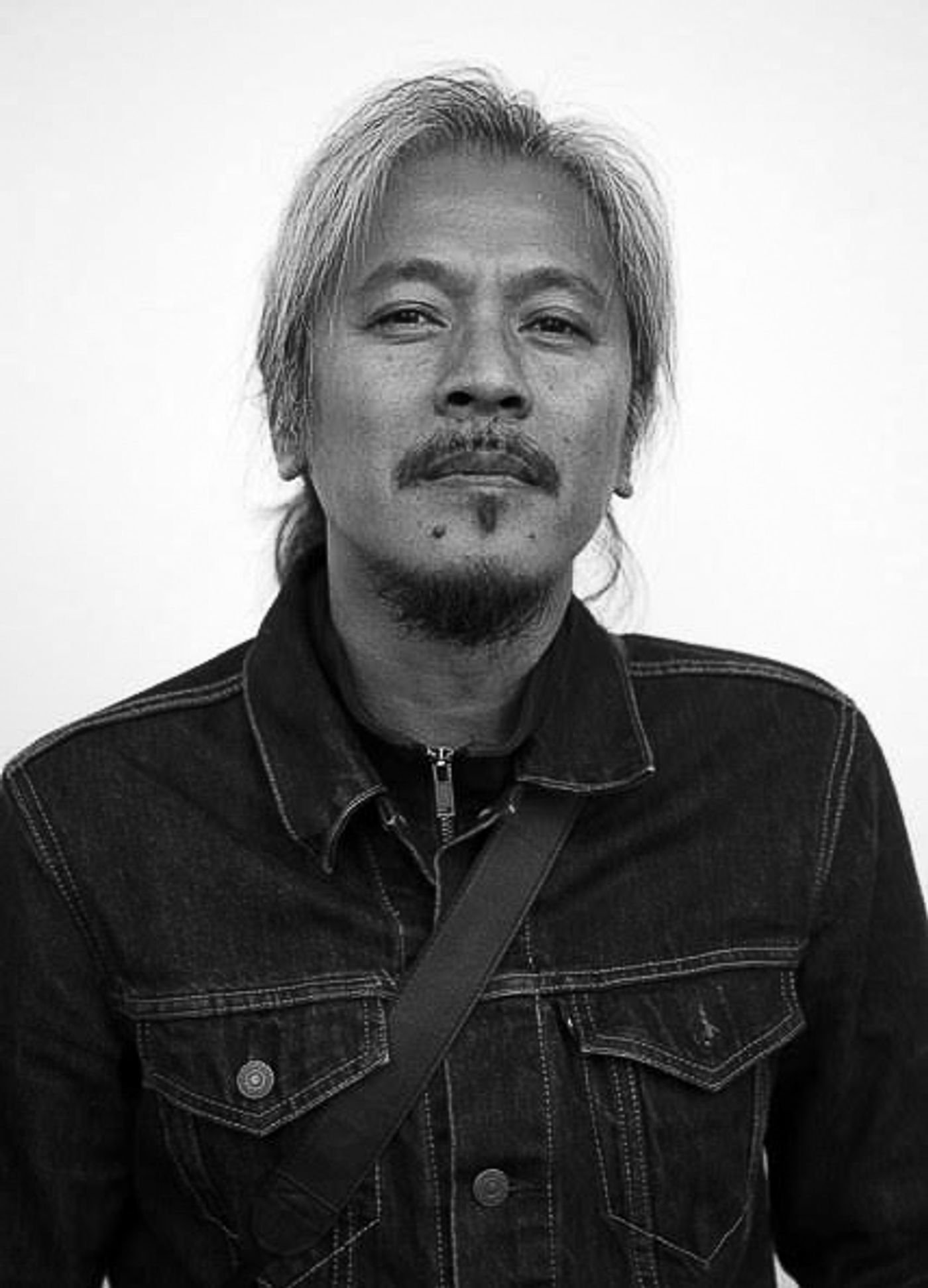 Lav Diaz in a denim jacket, long grey hair and a goatee, looking straight at the camera