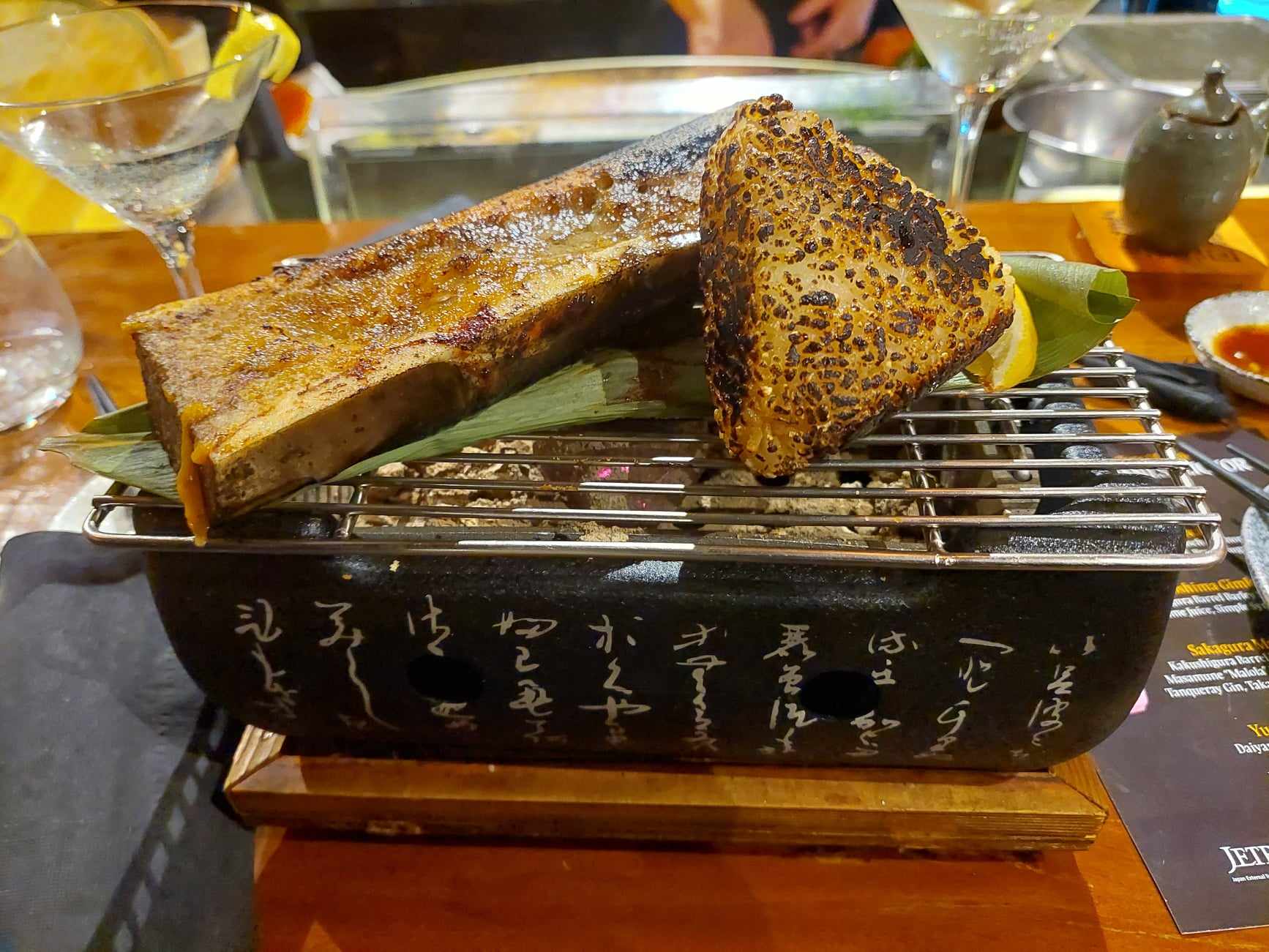 A split roasted bone marrow and a charred miso-glazed piece on a tabletop grill over a black brazier inscribed with Japanese calligraphy