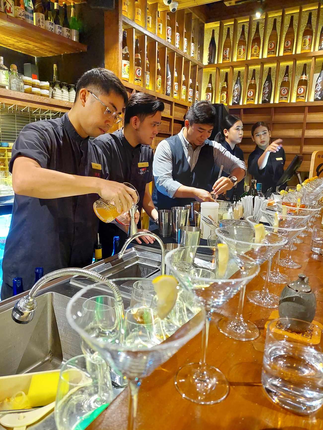 Bartenders in black mix cocktails behind a bar lined with martini glasses, a wall of shochu and sake bottles glowing behind them