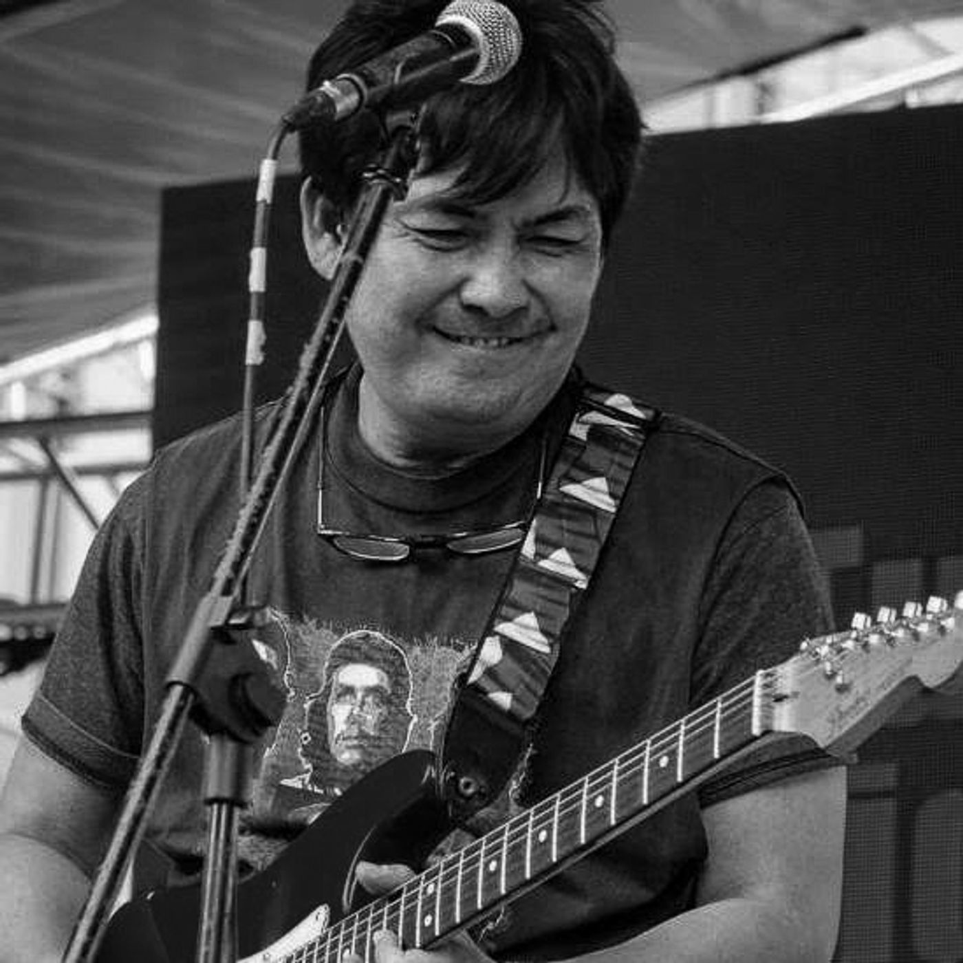Chickoy Pura laughing with his eyes shut as he plays a Stratocaster onstage, in black and white