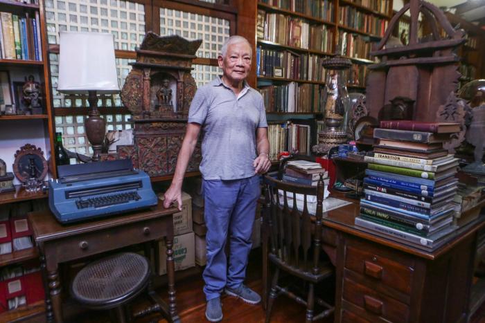 Dr. Jaime Laya stands between a blue electric typewriter and a stack of art books, in a library of carved santos and floor-to-ceiling shelves