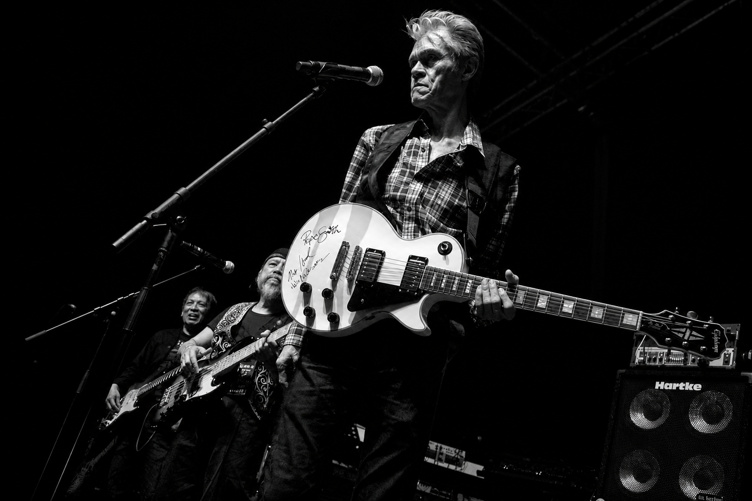 Pepe Smith onstage in a plaid shirt with a white Les Paul, two bandmates and a Hartke bass stack behind him in the dark
