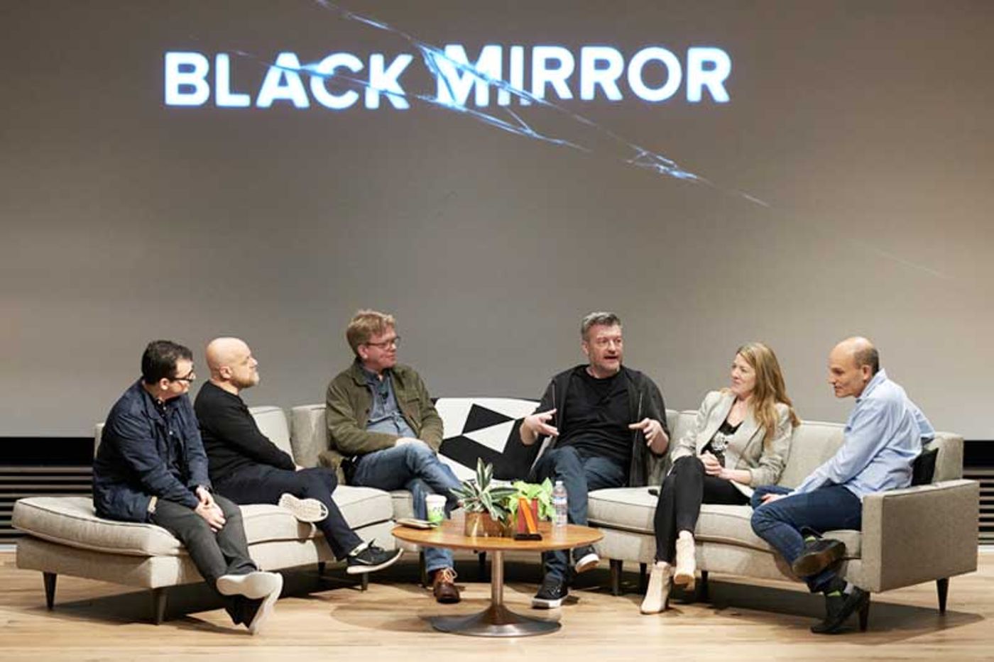 Charlie Brooker and five Netflix panelists chat on couches beneath a Black Mirror projection