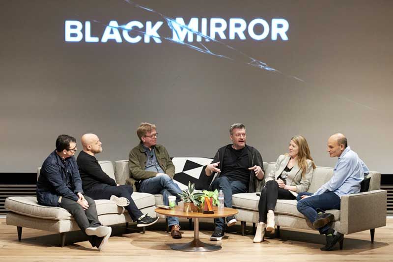 Charlie Brooker and five Netflix panelists chat on couches beneath a Black Mirror projection