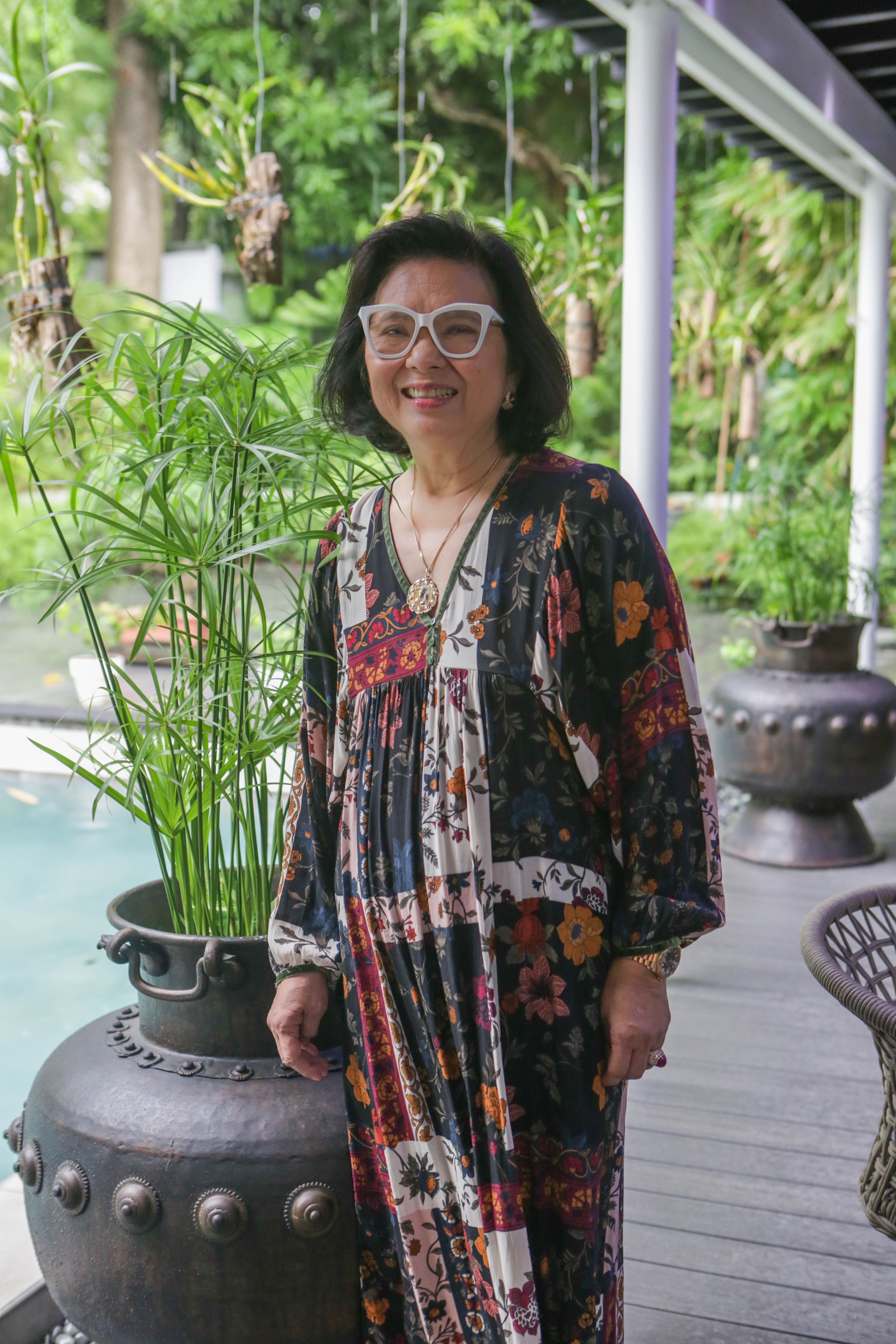 Menchu Tantoco-Lopez in a patterned dress smiling beside a papyrus plant in a studded metal urn, the swimming pool and garden behind her