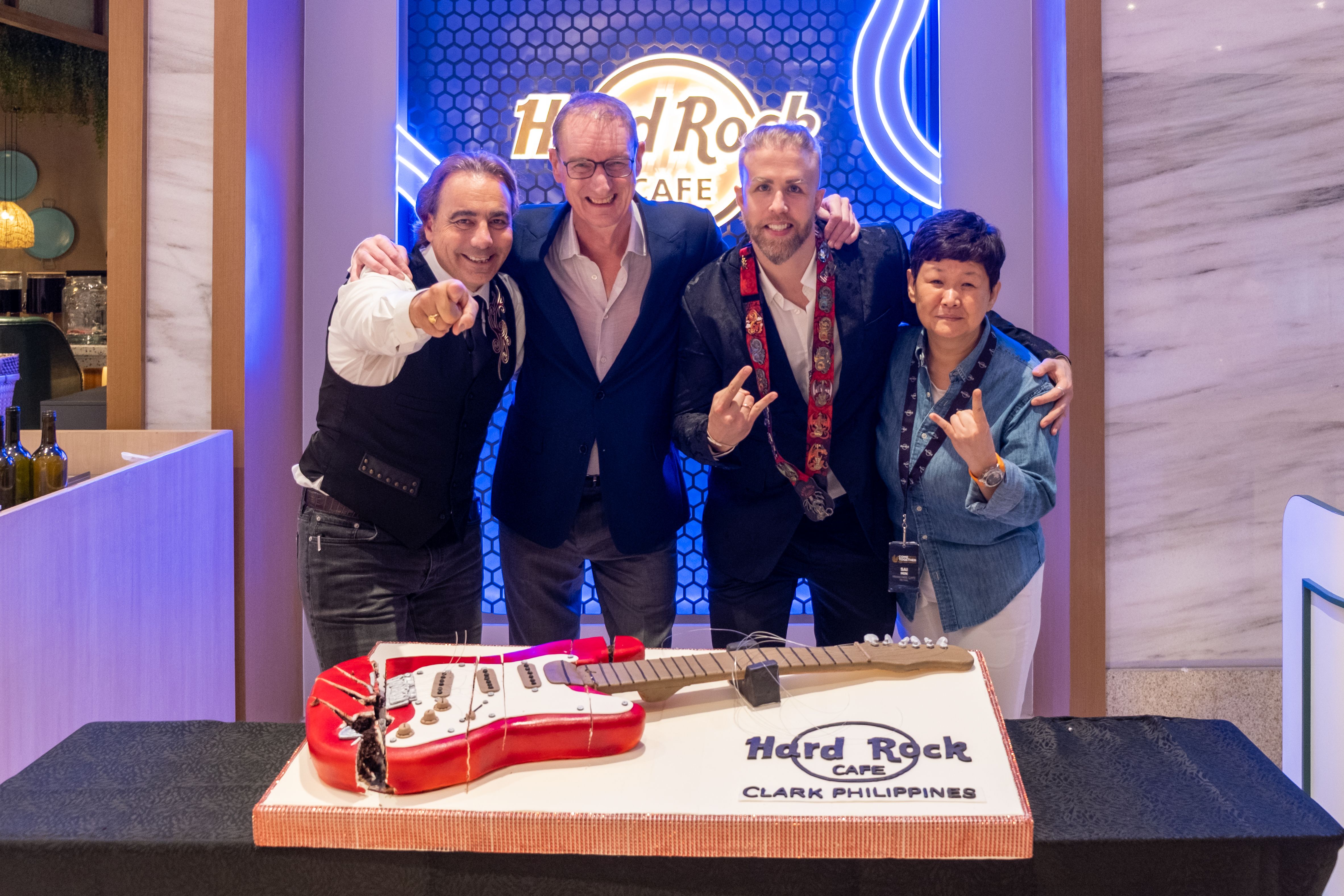 Four smiling guests pose arm in arm behind the guitar-shaped cake under the blue LED wall