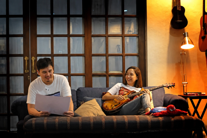 Martin Honasan and Barbie Almalbis on a sofa in a warm-lit room, he looking through a large sketchpad, she playing a semi-hollow electric guitar, guitars hanging on the wall behind