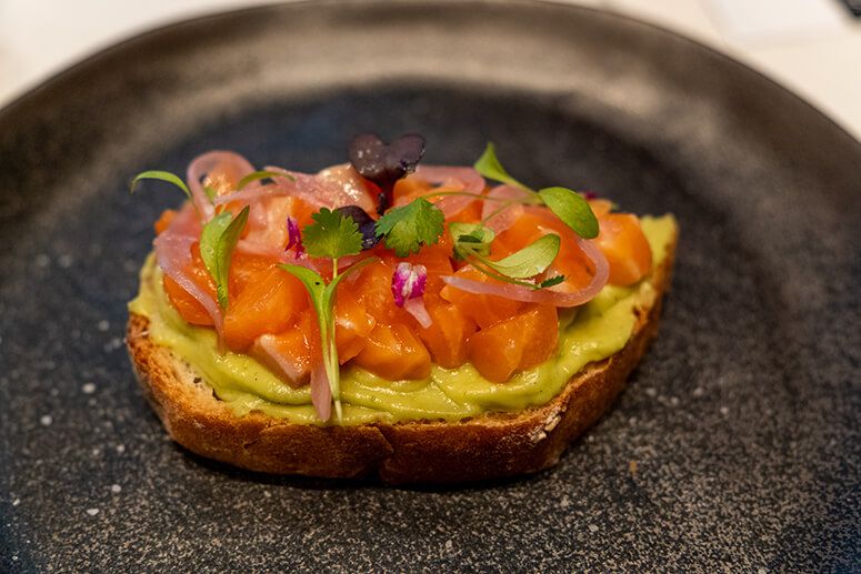 Salmon and avocado toast topped with diced salmon, pickled red onions and edible flowers on a black plate