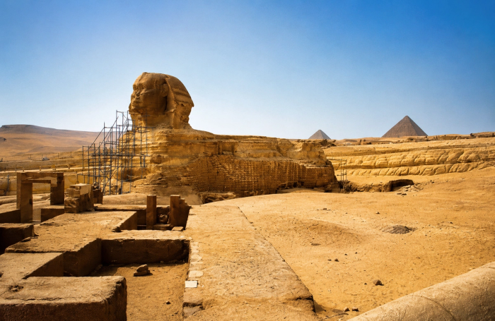 The Great Sphinx under scaffolding at Giza, with two pyramids rising in the distance