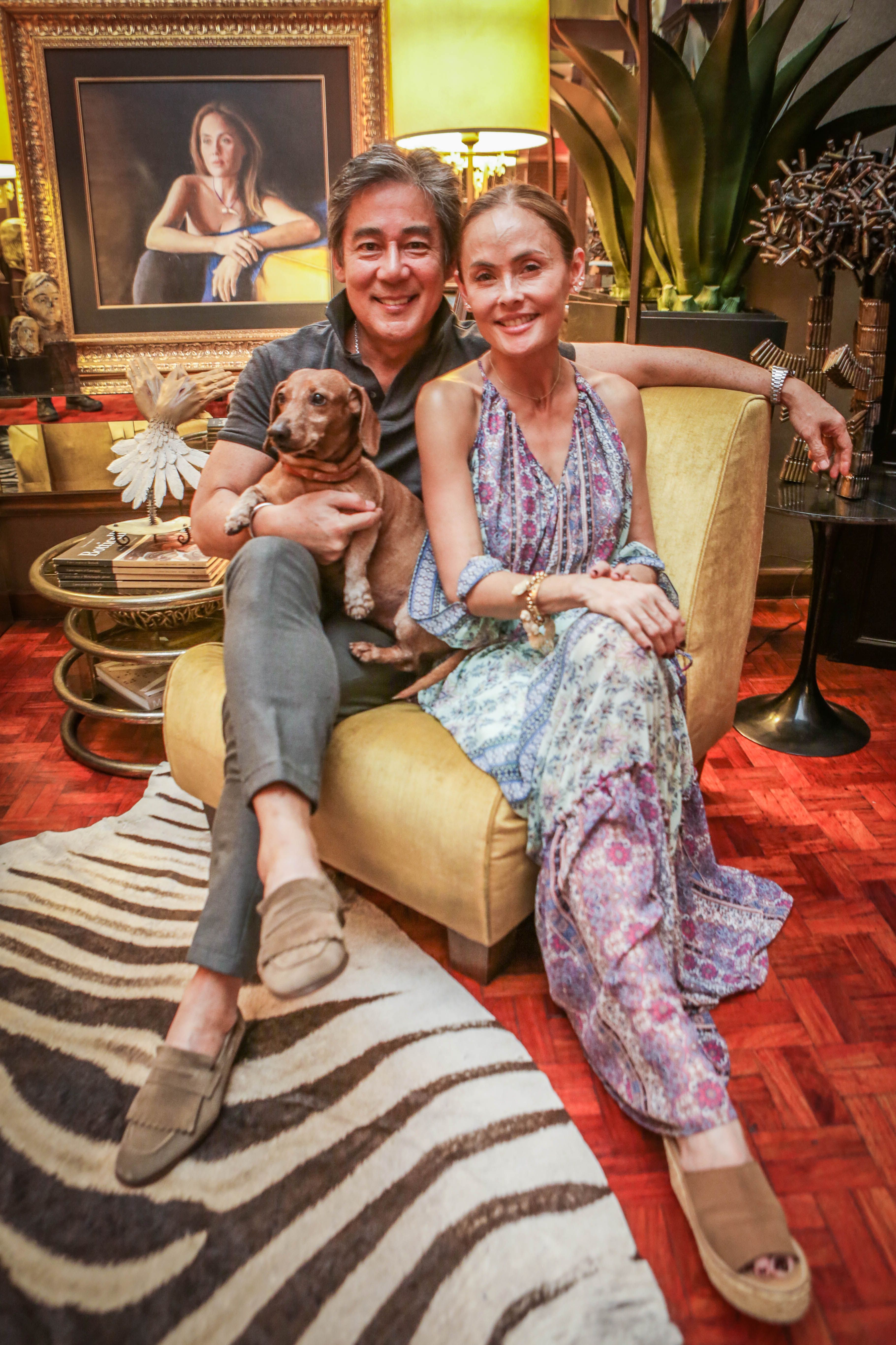 Raul Francisco and Joanna Preysler-Francisco sit close on a mustard sofa holding their brown dachshund, a framed portrait and gilded lamp behind them