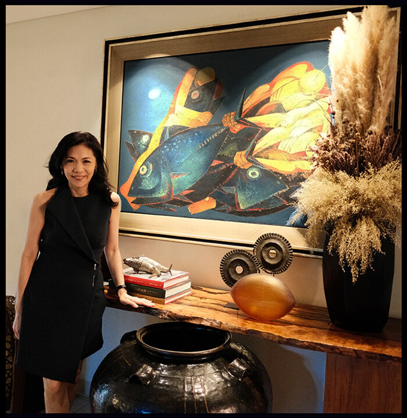 Kaye Tinga standing in a black dress next to a rustic wooden console table, with a large Ang Kiukok painting of three yellow and blue fish hanging behind her