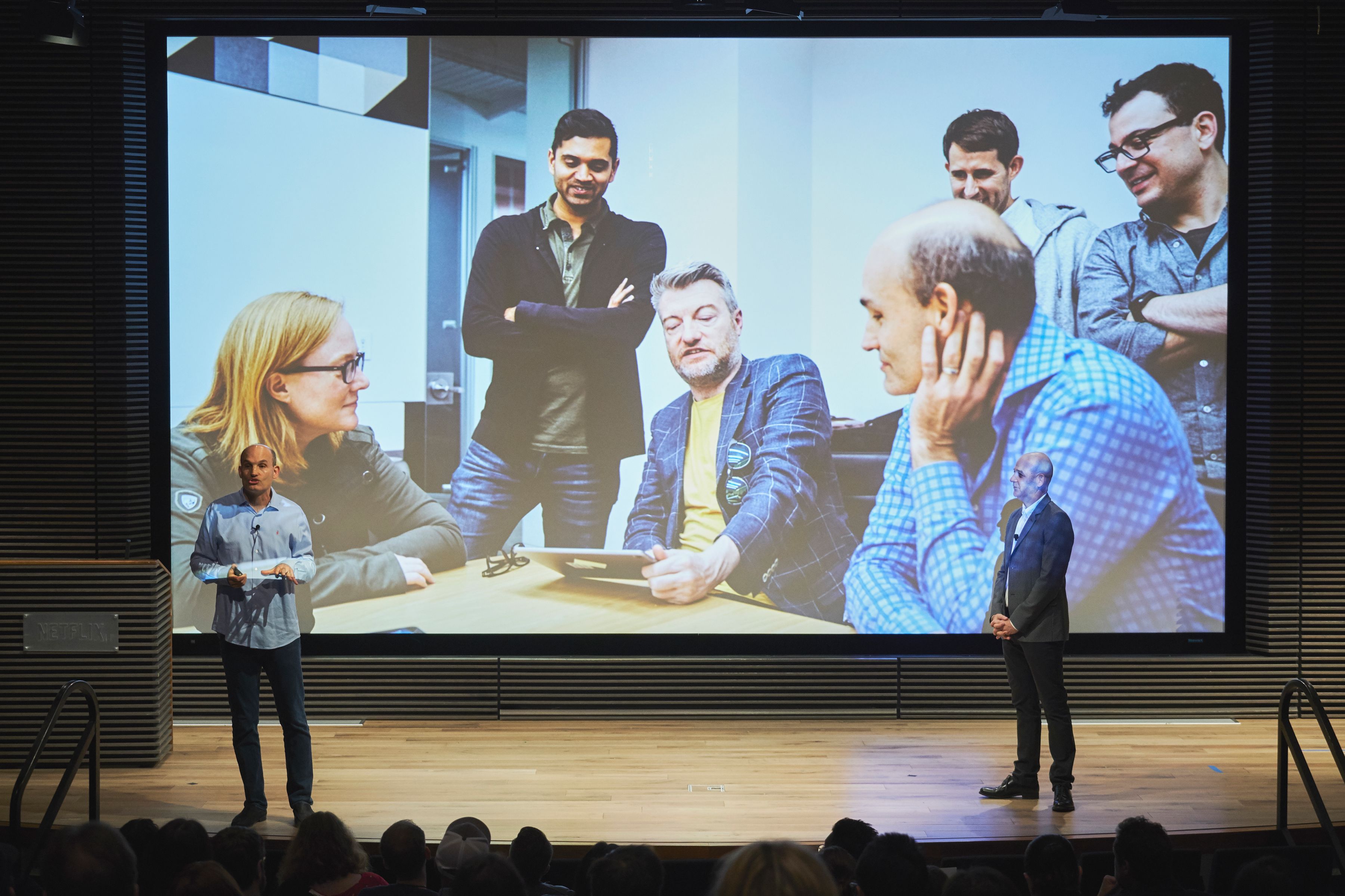 Two speakers onstage in front of a large photo of the Black Mirror team gathered around a table