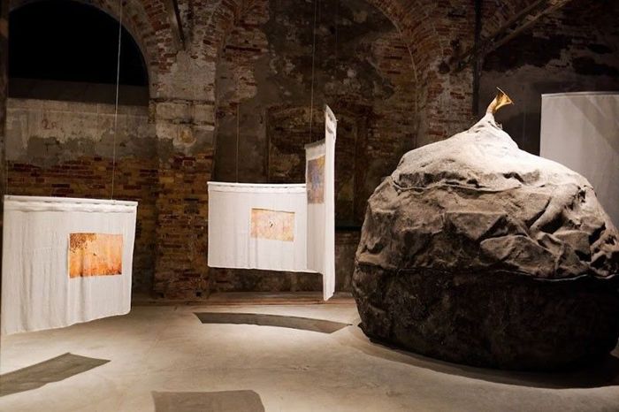 Installation inside the Philippine Pavilion at the Venice Biennale featuring a large grey boulder with a brass horn sticking out and hanging white textile sheets