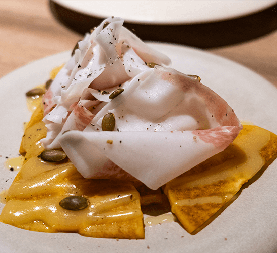 Translucent ribbons of lardo draped over golden persimmon slices scattered with pumpkin seeds